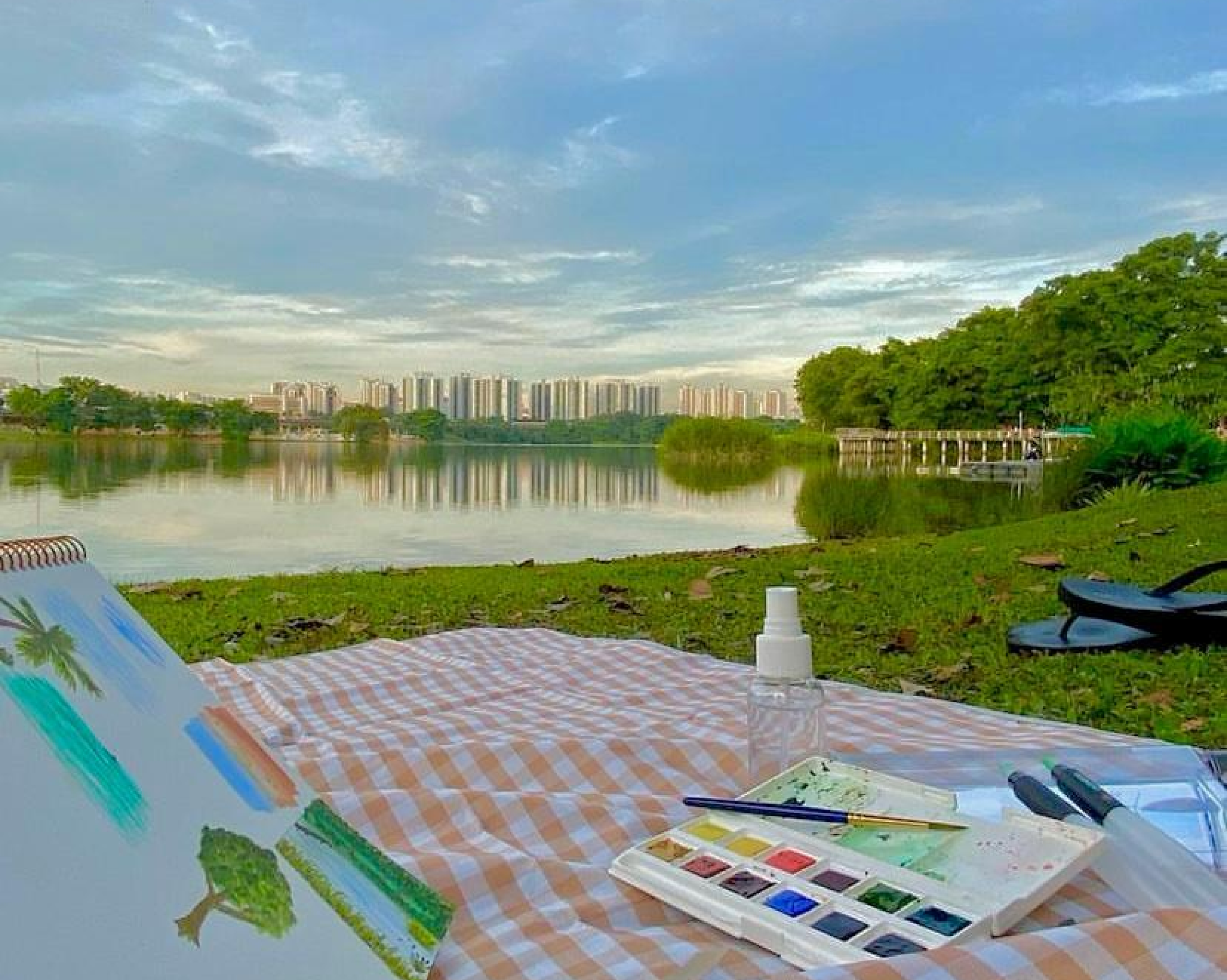 Sketchbook and watercolour paints on a picnic mat, looking out onto a reservoir